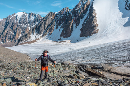 Hiker with trekking poles walks near large glacier tongue in high mountains. Scenic landscape with tourist near glacier in sunlight. Man with sunglasses and camera among snow mountains in sunny day.の写真素材
