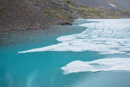 Atmospheric mountain landscape with frozen alpine lake during snowfall. Clear ice floats on water surface of lake. Awesome mountain scenery with icy mountain lake. Flakes of snow on water background.の写真素材