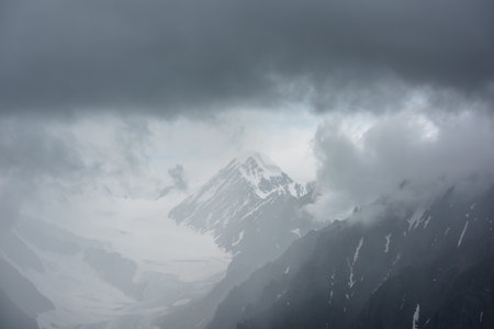 Atmospheric landscape with fuzzy silhouettes of sharp rocks and snowy mountain top in low clouds during rain. Dramatic view to large snow mountains and glacier blurred in rain haze in gray low clouds.の写真素材