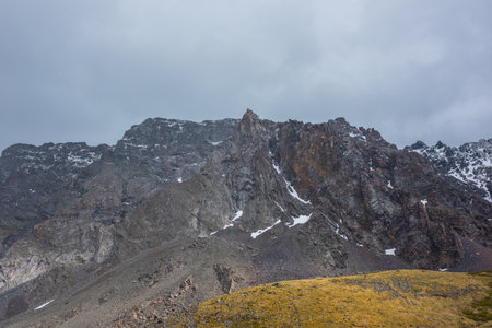Dramatic mountain landscape with sunlit golden hill and sharp pinnacle on high mountain range in cloudy sky during snowfall. Colorful alpine scenery with peaked mountain top at changeable weather.の写真素材