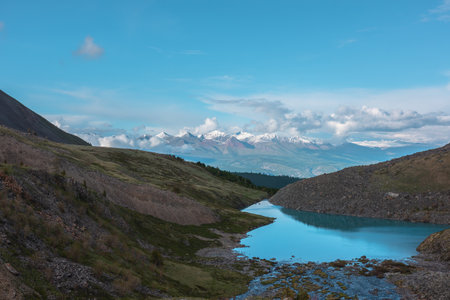 Atmospheric landscape with azure mountain lake against high snow-white mountain range in sunlight in low clouds. Dramatic evening view to alpine lake and large sunlit snow mountains in low clouds.の写真素材