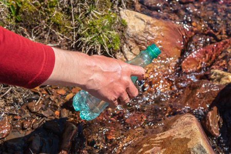 Man replenishes water from mountain stream in bright sun. Tourist fills bottle with mineral water in sunny day. Hand with bottle in clear water stream in sunlight. Replenishment of water during hike.の写真素材
