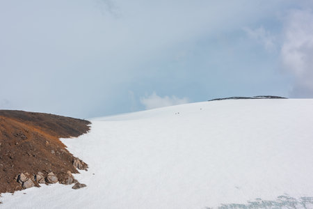 Scenic alpine landscape with small people on high snowy mountain in sunlight under clouds in blue sky. Awesome mountain scenery with small tourists on sunlit large snow mountain at changeable weather.の写真素材