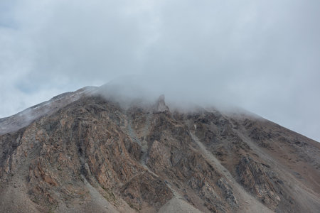 Dramatic mountain landscape with sharp rocks in rainy low clouds. Beautiful alpine scenery with large rocky mountain with snow top in low gray cloudy sky. Gloomy gray rainy weather in high mountains.の写真素材