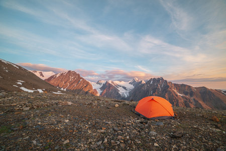 Awesome landscape with vivid orange tent with view to high snow mountains in sunrise colors. Colorful scenery with sunlit gold rocks and mountains at sunrise. Gold early morning at very high altitude.の写真素材