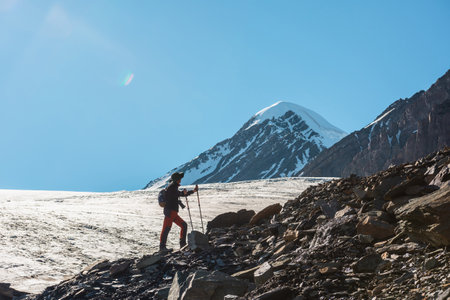 Scenic alpine landscape with silhouette of hiker with trekking poles against large glacier tongue and snow mountain peak in sunlight. Man with backpack in high mountains under blue sky in sunny day.の写真素材