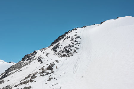 Climbers footprints on steep snowy slope of high snow mountain with rocks on top. Snow-white mountain under blue sky. Minimal landscape with snowy mountain with alpinists footprints at high altitude.の写真素材