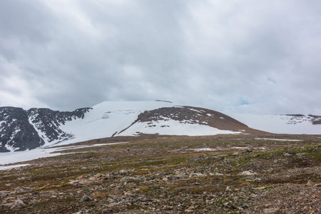 Dramatic landscape with stone field and large snowy mountain dome under gray cloudy sky. Atmospheric scenery with high snow mountain in dome shape in center at cloudy weather. Overcast in mountains.の写真素材