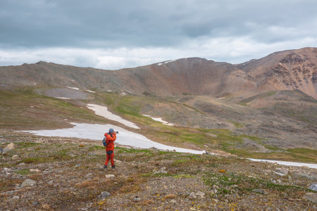 Atmospheric scenery with man in red against mountain range with sharp rocks under gray cloudy sky. Tourist with camera shoots dramatic mountain landscape. Man photographs mountains at gloomy weather.の写真素材