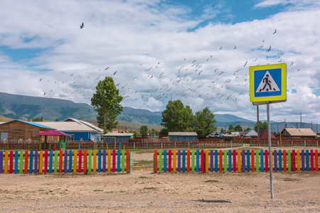 Flock of birds flies over colored playground at changeable weather. Dramatic landscape with sunlit empty playground behind multicolor fence in mountain village against high mountain range under cloudsの写真素材