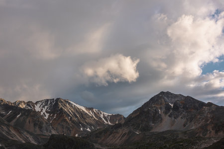Dramatic mountain landscape with high mountain range with sharp rocky pinnacle under clouds of sunset color in gloomy sky. Dark atmospheric scenery with large mountains with snow at changeable weatherの写真素材