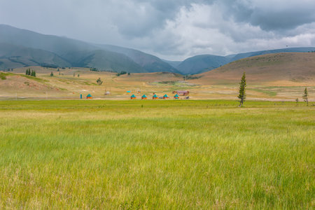 Scenic view from sunlit grassy steppe to small houses on background of green high mountain range in rainy clouds. Dramatic landscape with meadow and small lodges in mountains at changeable weather.の写真素材