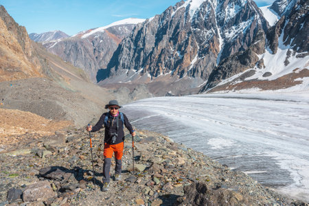 Hiker with trekking poles walks near large glacier tongue in high mountains. Scenic landscape with tourist near glacier in sunlight. Man with sunglasses and camera among snow mountains in sunny day.の写真素材