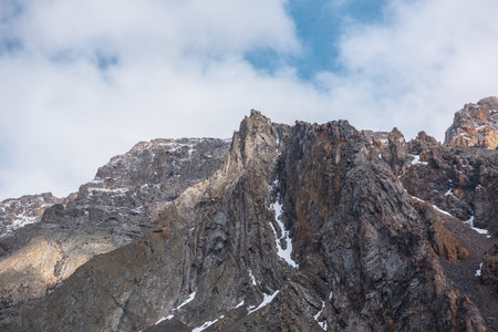 Atmospheric landscape with sharp pinnacle on sunlit high mountain range under clouds in blue sky. Beautiful alpine scenery with peaked mountain top in sunlight under cloudy sky at changeable weather.の写真素材