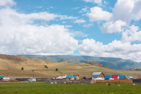 Dramatic alpine landscape with small houses in sunlit green meadow against high mountain range under clouds in blue sky. Beautiful mountain village at changeable weather. Scenic view to high mountainsの写真素材