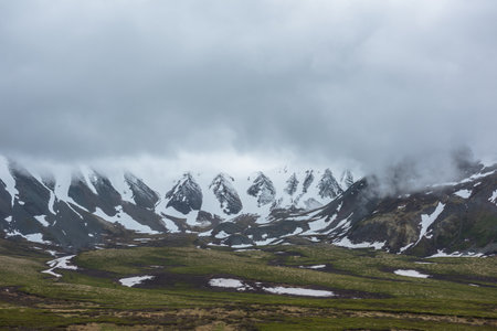 Dramatic alpine landscape with snowy mountains in gray low clouds. Bleak atmospheric scenery of tundra under lead gray sky. Gloomy minimalist view to mountain range among low rainy clouds in overcast.の写真素材