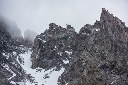 Gloomy mountain landscape with high sharp rockies with snow in gray low clouds. Closeup of sharp rocks and peaked tops in gray cloudy sky. Bleak overcast scenery with mountain range with pointy rocks.の写真素材