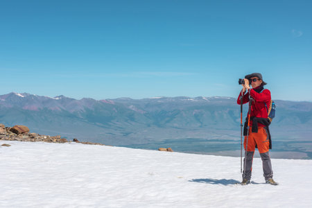 Tourist in red walks on snow mountain near abyss edge on high altitude under blue sky in sunny day. Man with camera on snowy mountain near precipice edge with view to large mountain range in away.の写真素材