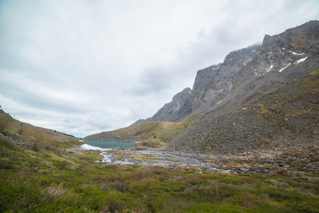 Atmospheric landscape with azure mountain lake in green valley and rocky mountain range with peaked top in low clouds. Beautiful alpine lake and high sharp rocks under cloudy sky at gloomy weather.の写真素材
