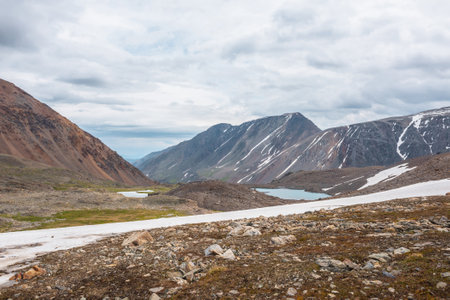Dramatic landscape with glacier and stone hill between two mountain lakes in sunlight under cloudy sky. Two small lake in high mountain valley with view to mountain vastness at changeable weather.の写真素材