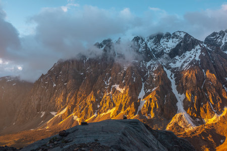 Scenic landscape with snowy mountain top in low clouds in golden sunrise colors. Colorful view to snow mountains and rocks in gold morning sunlight in low clouds. Awesome scenery with golden rocks.の写真素材
