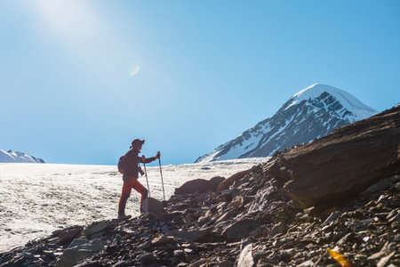 Scenic alpine landscape with silhouette of hiker with trekking poles against large glacier tongue and snow mountain peak in sunlight. Man with backpack in high mountains under blue sky in sunny day.の写真素材