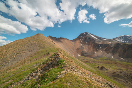 Scenic landscape with sunlit green grassy stone hill and high orange mountain ridge under white clouds in blue sky. Colorful scenery with rocky mountains and sharp rocks in sunlight under cloudy sky.の写真素材