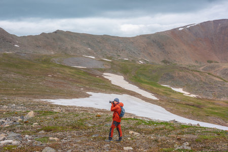 Atmospheric scenery with man in red against mountain range with sharp rocks under gray cloudy sky. Tourist with camera shoots dramatic mountain landscape. Man photographs mountains at gloomy weather.の写真素材