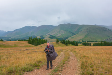 Satisfied tourist returns from mountains in overcast. Happy man in raincoat walks through hills and forest in bad weather. Traveler goes towards adventure. Hiker and mountain range under cloudy sky.の写真素材