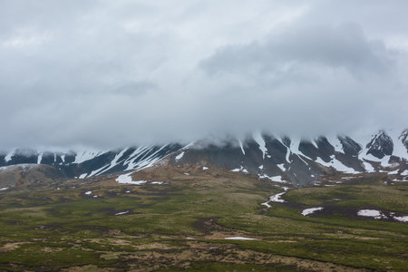 Dramatic alpine landscape with snowy mountains in gray low clouds. Bleak atmospheric scenery of tundra under lead gray sky. Gloomy minimalist view to mountain range among low rainy clouds in overcast.の写真素材