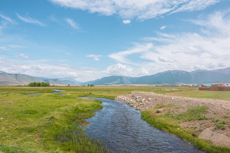 Scenic green landscape with mountain creek in sunlit grassy steppe among mountains under clouds in blue sky at changeable weather. Colorful scenery with mountain brook with clear water in bright sun.の写真素材