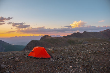 Scenic alpine landscape with tent at very high altitude with view to large mountains in orange dawn sky. Vivid orange tent with awesome view to high mountain range under cloudy sky in sunset colors.の写真素材