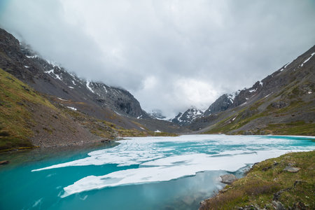 Atmospheric mountain landscape with frozen alpine lake and high snowy mountains. Awesome overcast scenery with icy mountain lake on background of snow mountains in low clouds. Scenic view to ice lake.の写真素材