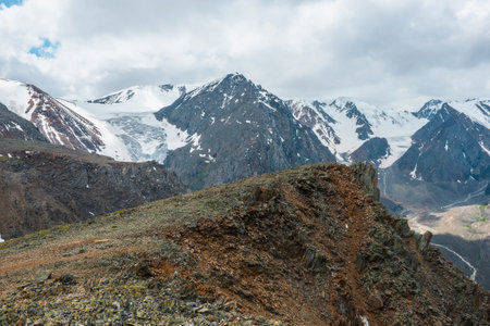 Vivid landscape near precipice edge with sharp rocks with view to large snowy mountains and glacier under cloudy sky. Picturesque view from stone hill to high mountain range with shadows from clouds.の写真素材