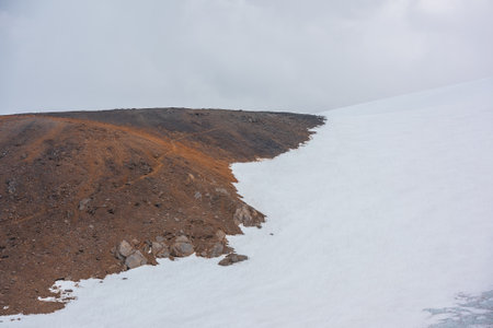 Scenic landscape with hiking trail on stone hillside to high snowy mountain in sunlight under gray cloudy sky. Tourist path on sunlit rocky mountainside to large snow mountain at changeable weather.の写真素材