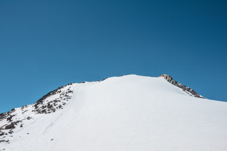 Climbers footprints on steep snowy slope of high snow mountain with rocks on top. Snow-white mountain under blue sky. Minimal landscape with snowy mountain with alpinists footprints at high altitude.の写真素材