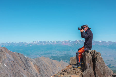 Tourist sitting on stone near abyss edge on high altitude under blue sky in sunny day. Man with camera on high rock near precipice edge with wonderful view from above to large mountain range in away.の写真素材