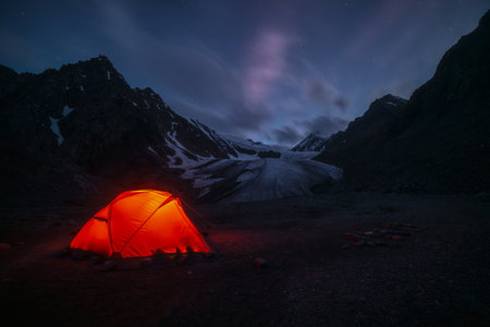 Awesome mountain landscape with vivid orange tent near large glacier tongue under clouds in night starry sky. Tent glow by orange light with view to glacier and mountains silhouettes in starry night.の写真素材