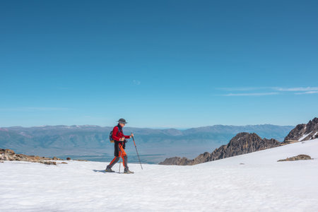 Tourist in red walks on snow mountain near abyss edge on high altitude under blue sky in sunny day. Man with camera on snowy mountain near precipice edge with view to large mountain range in away.の写真素材