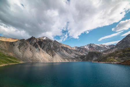 Deep mountain lake of phantom blue color among high mountains with pointed peak in changeable weather. Wonderful dramatic view to deep blue mountain lake among sunlit sharp rocks under cloudy sky.の写真素材