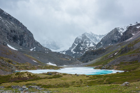 Atmospheric mountain landscape with frozen alpine lake and high snowy mountains. Awesome overcast scenery with icy mountain lake on background of snow mountains in low clouds. Scenic view to ice lake.の写真素材