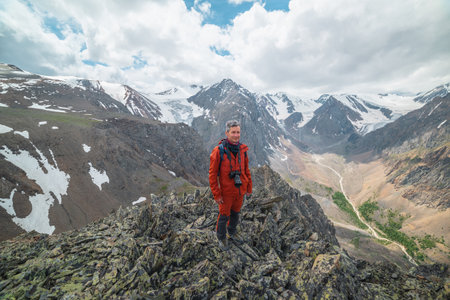 One man in red with camera near precipice edge with sharp rocks with view to large snowy mountains and glacier under cloudy sky. Tourist on stone hill with view to mountain range at changeable weatherの写真素材