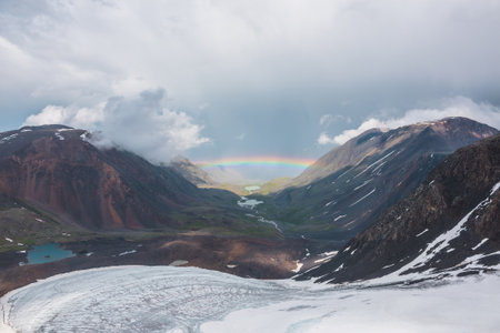 Bleak landscape with vivid rainbow above mountain lake. Gloomy scenery with bright rainbow above glacier and glacial lake in mountain valley. Top view to colorful rainbow and low clouds in mountains.の写真素材