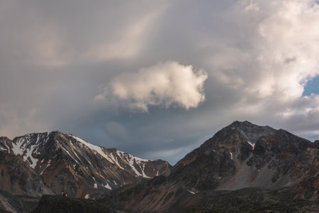 Dramatic mountain landscape with high mountain range with sharp rocky pinnacle under clouds of sunset color in gloomy sky. Dark atmospheric scenery with large mountains with snow at changeable weatherの写真素材