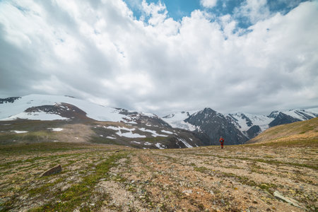 One man walks with view to high snowy mountain range with sharp tops and glaciers under gray cloudy sky. Colorful landscape with tourist on background of large snow mountains at changeable weather.の写真素材