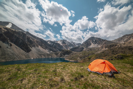 Vivid orange tent on grassy hill with view to mountain lake of phantom blue color among high mountains in changeable weather. Tent near deep blue lake among sunlit large mountains under cloudy sky.の写真素材