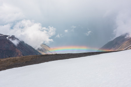 Minimalist alpine landscape with vivid rainbow. Scenic minimal view from snow hill to bright rainbow in mountain valley. Top view from snow mountain to colorful rainbow and low clouds in rainy weatherの写真素材