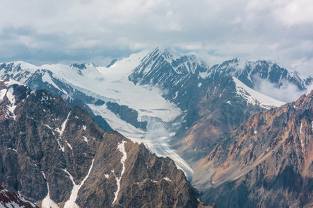 Dramatic landscape with long glacier tongue and high snow mountains under cloudy sky. Wonderful aerial view to sunlit large glacier and snowy mountain peak in clouds. Top view to majestic mountains.の写真素材