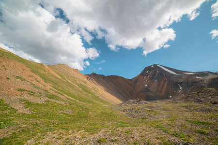 Scenic landscape with sunlit green grassy stone hill and high orange mountain ridge under white clouds in blue sky. Colorful scenery with rocky mountains and sharp rocks in sunlight under cloudy sky.の写真素材