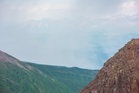 Scenic aerial view from high sharp rocks to green hills and sunlit mountain range in rainy clouds during rain. Dramatic atmospheric landscape with mountain vastness in low clouds in changeable weatherの写真素材